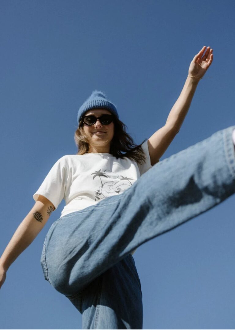 Woman in a blue beanie and sunglasses kicking one leg up against a blue sky