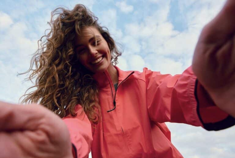 Smiling woman in a pink jacket reaching toward the camera under a blue sky
